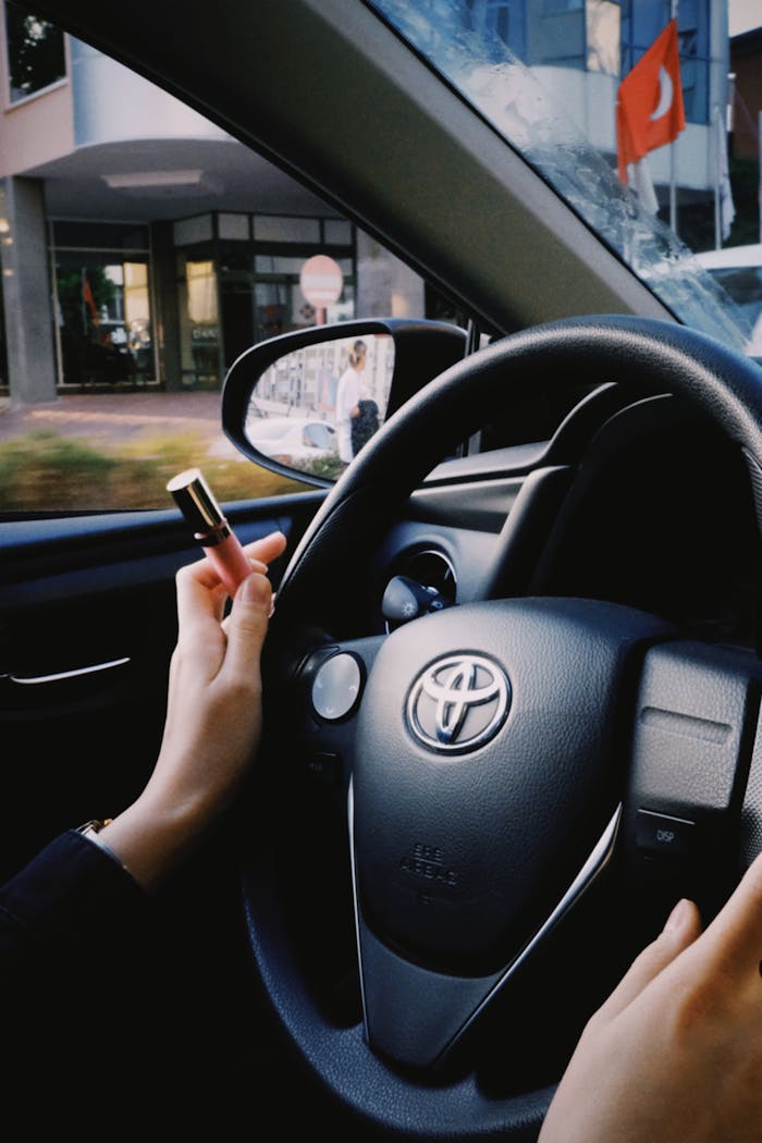 Hands holding lipstick while steering a Toyota car, showing urban scenery in the background.