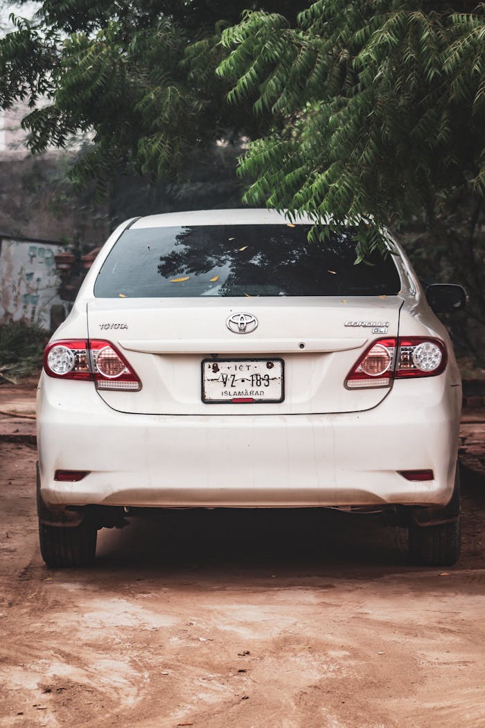A white sedan with license plate parked under trees on a dirt road, captured outdoors.